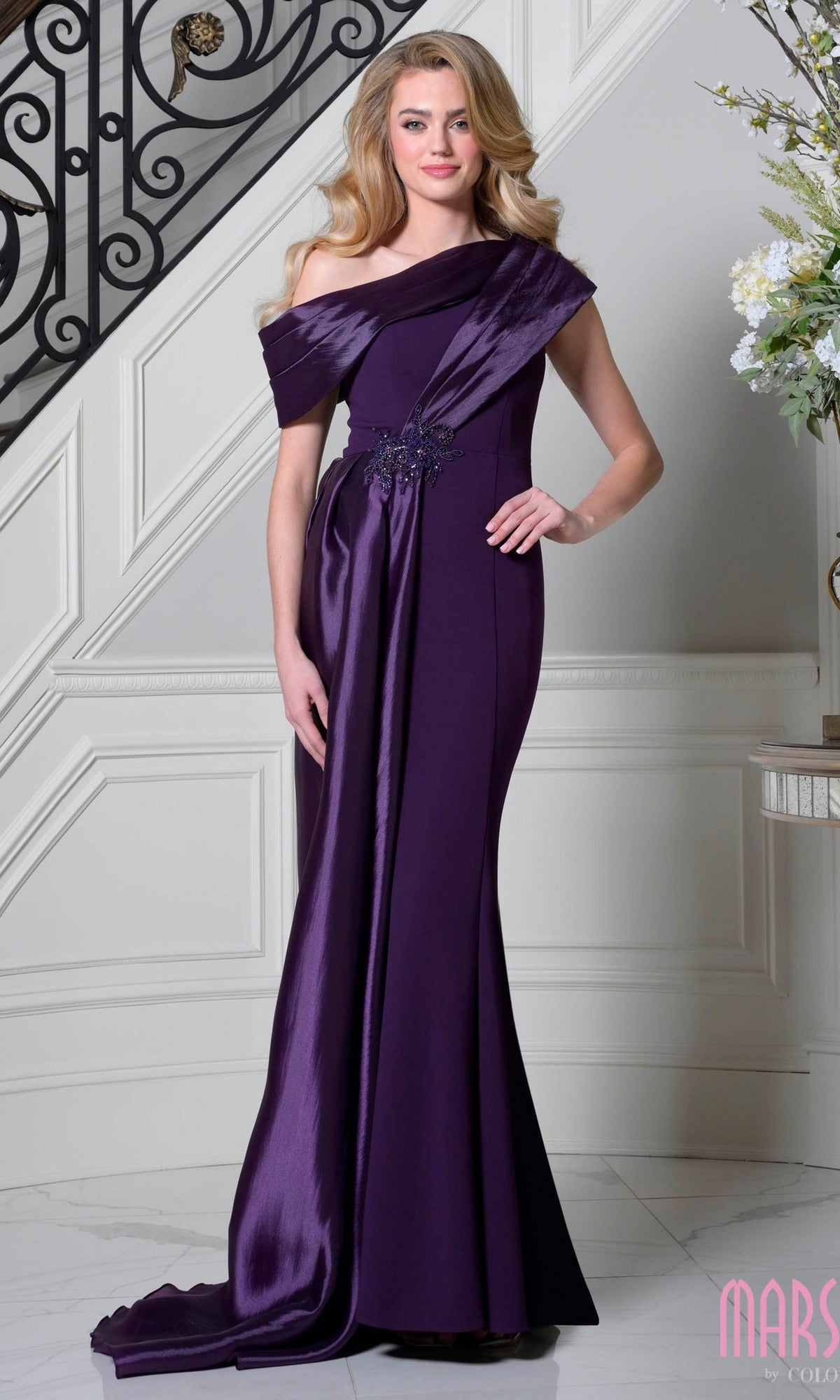 A woman poses gracefully indoors by a staircase in the Marsoni M346 Asymmetrical Long Formal Dress, an off-the-shoulder, floor-length gown in elegant eggplant with draped satin and floral embellishments.